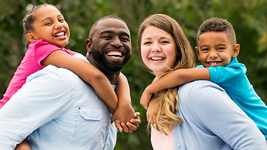 A family poses for a photo outdoors. The mom and dad are giving the kids a piggy-back ride.
